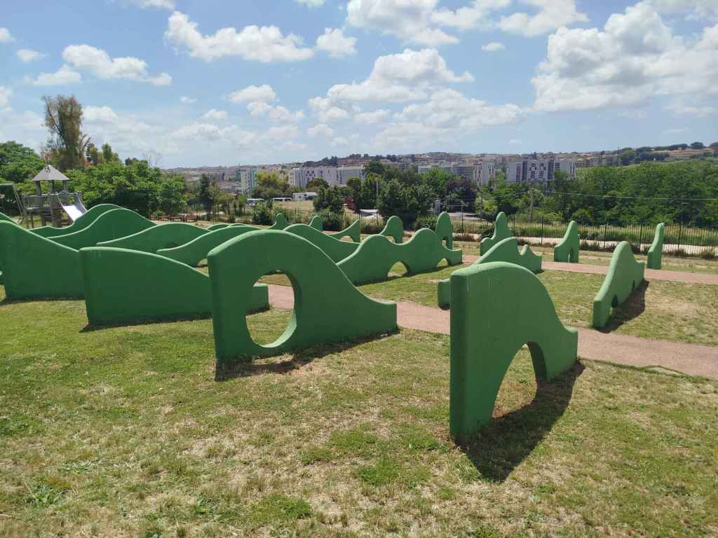 A children's playground featuring green textured structures resembling hills or waves, set against a clear blue sky with fluffy clouds.
