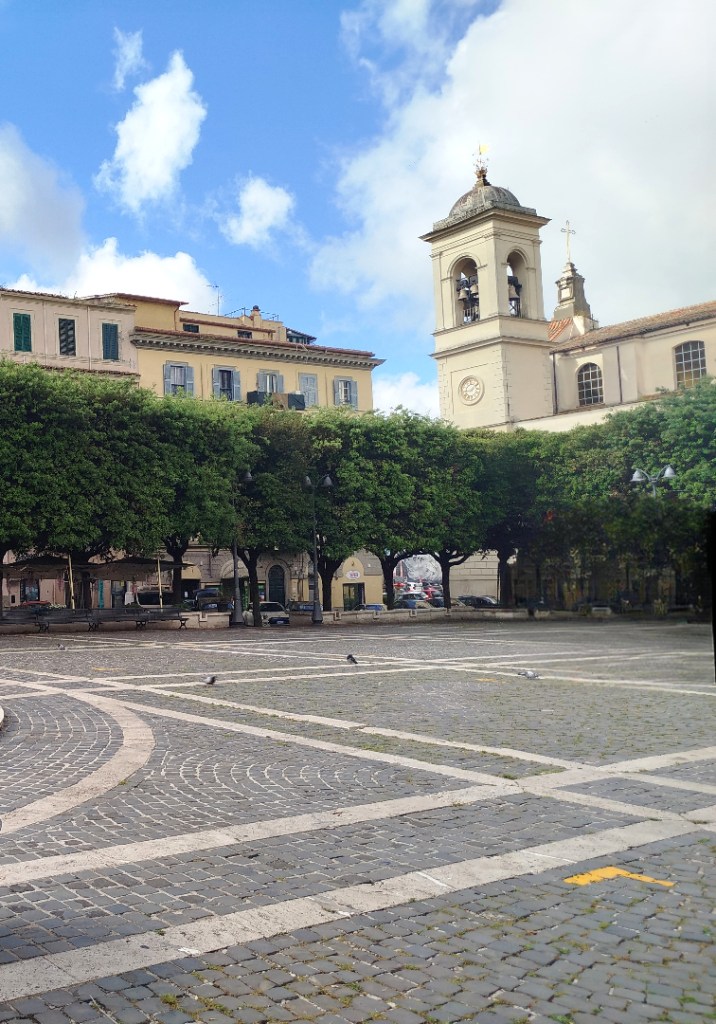 A scenic view of a plaza featuring stone pavement, greenery, and historical buildings, including a clock tower, under a partly cloudy sky.